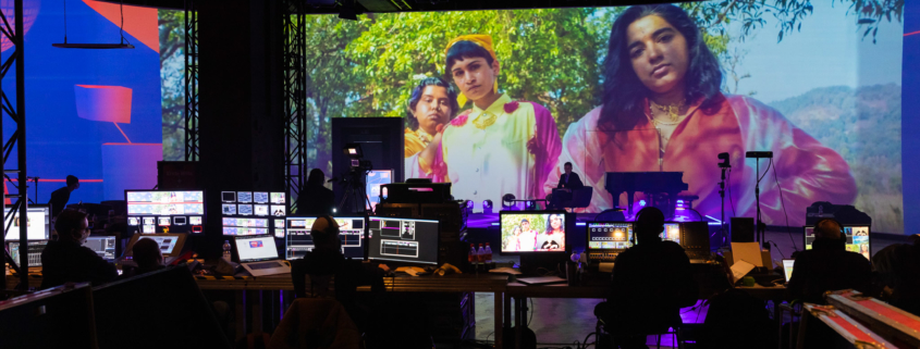 Produzent:innen sitzen vor einer Vielzahl an Bildschirme vor eine Großen Leinwand im Rahmen der Produktion der Most Wanted: Music Konferenz 2020